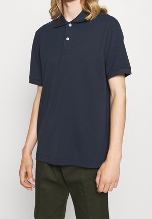 Person iført en navyblå polo med korte ærmer og to knapper samt olivengrønne bukser, stående mod en ensfarvet baggrund.