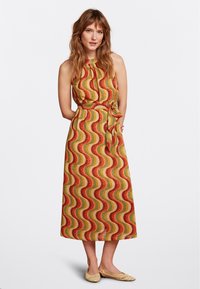 Mehrfarbiges, wellenmusterndes Kleid in Gelb, Orange und Grün. Trägerschnitt, mit Gürtel in der Taille und knielang. Getragen mit flachen geflochtenen Schuhen.