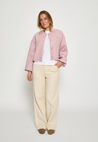 Ružová, textúrovaná skrátená bunda s gombíkmi, kombinovaná s bielou košeľou a béžovými širokými nohavicami. Hnedé topánky dotvárajú outfit.