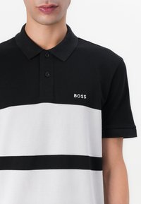 Svart og hvit polo skjorte med krage, som har en horisontal svart stripe og ordet "BOSS" brodert i hvitt på venstre bryst.