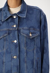 Jeans jakna v temno modri barvi z diskretnim krožnim vzorcem. Ima srebrne kovinske gumbke, dve žepki na prsih in kontrastno šivanje.