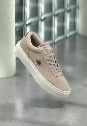 Lys beige semsket skinnsko med hvit såle, snøring, og svart fuglelogo på siden, vist mot en moderne bakgrunn av glassblokker.