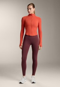 Rood nauwsluitend shirt met lange mouwen, hoge kraag en volledige ritssluiting, gecombineerd met donkerrode legging met een accentstreep aan de zijkant; textuurstof.