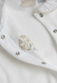 Bodysuit bébé blanc avec un motif floral brodé en beige doux et vert. Doté de boutons-pression métalliques et d'un col froncé. Texture de tissu douce.