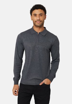 Uomo che indossa un maglione grigio scuro a maniche lunghe con collo a mezza zip, in piedi con una mano in tasca davanti a uno sfondo chiaro e uniforme.