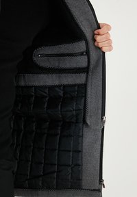 Chaqueta de mezcla de lana gris sin cremallera, que revela un forro acolchado negro. Sus características incluyen una textura suave y costuras contrastantes.