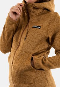 Chaqueta de forro polar marrón con capucha, cierre de cremallera, bolsillos tipo canguro y un parche de logo negro. Presenta un tejido texturizado y un ribete en contraste.