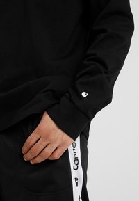 Sort langærmet skjorte med et lille hvidt logo på manchetten, parret med sorte bukser med en hvid sidestribe med branding.