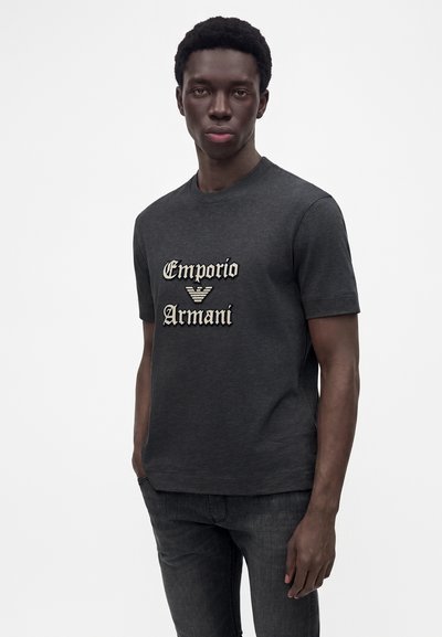 Νέος άνδρας που φοράει ένα σκουρογκρι t-shirt Emporio Armani και μαύρα τζιν, στέκεται μπροστά σε λευκό φόντο.