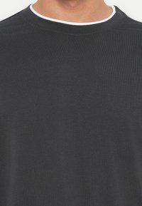 Close-up van een persoon die een donkergrijs T-shirt met een witte rand rond de kraag draagt, tegen een effen achtergrond.