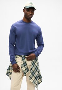 Modra dukserica s okruglim izrezom i logotipom, uparena s bež hlačama. Zeleno-bjela košulja je vezana oko struka. Svijetla pozadina.