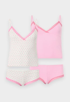 Mujer de pie descalza con camisola rosa y pantalones cortos a juego con encaje en un fondo claro y liso.