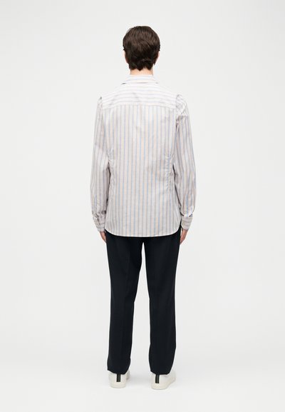 Άνδρας που φοράει ανοιχτόχρωμο ριγέ πουκάμισο με μακριά μανίκια, μαύρο παντελόνι και λευκά αθλητικά παπούτσια, στέκεται με την πλάτη γυρισμένη σε απλό φόντο.