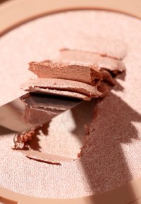 Illuminante in polvere compatta di colore rosa tenue, con una finitura luminosa. Un coltello metallico solleva sottili strati, rivelando una superficie liscia e testurizzata.