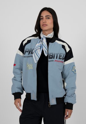 Sieviete, tērpusies zilā un melnā sacīkšu stila jakā ar uzrakstu "PROHIBITED RACING", rakstainu šalli, melnās biksēs un zelta gredzeniem.