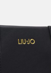 Gros plan d'une surface en cuir noir avec texte embossé doré "LIU JO" sur un produit, probablement un sac à main ou un portefeuille.