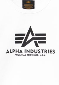 Witte sweatshirt met ronde hals, featuring een zwart logo met een gestileerde "A" en strepen. De tekst bevat "ALPHA INDUSTRIES" en "KNOXVILLE, TENNESSEE, U.S.A."