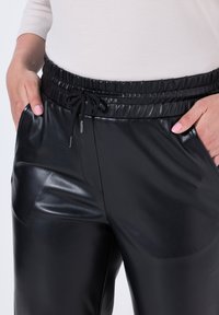 Černé kalhoty z umělé kůže s elastickým pasem, nastavovacím šňůrkovým pásem a bočními kapsami, s lesklou texturou a hladkým povrchem.