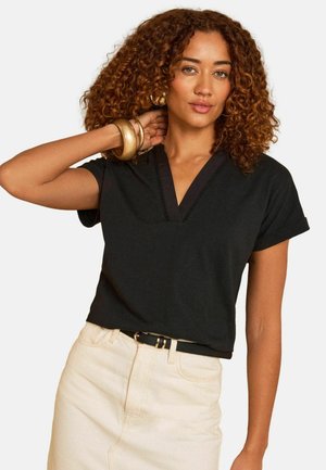 Nainen, jolla on kiharat hiukset ja joka pukeutuu mustaan V-kaula-aukkoinen lyhythihainen toppiin, beigen väriseen hameeseen, mustaan vyöhön, kultaisiin rengaskorvakoruihin ja rannekoruihin, poseeraa käsi kaulan takana.