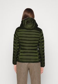 Donkergroene pufferjack met horizontale gestikte textuur, aangehechte capuchon en ritsluiting aan de voorkant. Gecombineerd met broek in een houndstoothpatroon.