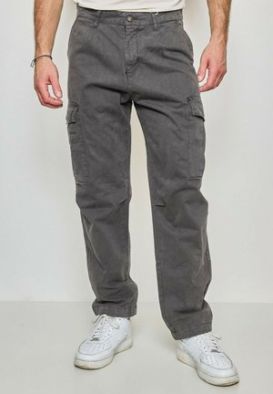 Homme portant un pantalon cargo gris foncé avec des poches latérales et des baskets blanches, debout contre un fond clair uni.