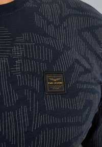 Camiseta texturizada azul marino con patrones lineales en gris claro. Incluye una etiqueta negra con letras doradas y un emblema.