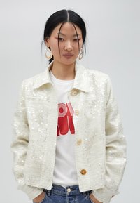 Paljettjacka i off-white, med krage, framknäppning och avslappnad passform. Bärs över en grafisk t-shirt och jeans, som visar upp den texturerade tyget.