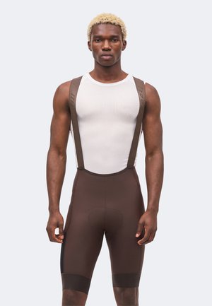 Urheilullinen mies, jolla on vaaleat hiukset, valkoinen hihaton verkkotoppi ja ruskeat pyöräilyhousut olkaimilla yksiväristä taustaa vasten.