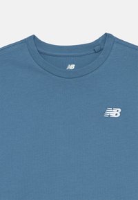 Modré tričko s kulatým výstřihem a bílými logy New Balance tištěnými uvnitř límce a na levé horní části hrudi.