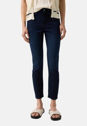 Kobieta ubrana w ciemnoniebieskie obcisłe dżinsy, kremowy kardigan i białe sandały z krzyżującymi się paskami, stojąca na jednolitym tle.