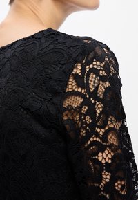 Crni čipkasti gornji dio s složenim cvjetnim uzorcima, prozirnim dugim rukavima i teksturiranim završetkom. Izrez je okrugao i elegantan.