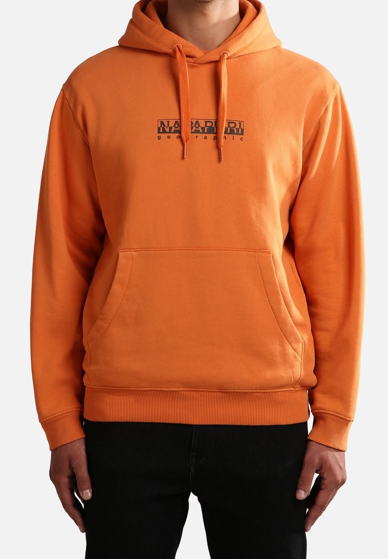 Homme portant un sweat à capuche orange avec poche avant et logo texte noir "Napapijri geographic" sur la poitrine, associé à un pantalon noir.
