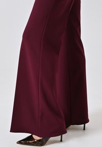 Pantaloni larghi color bordeaux che coprono tacchi alti a punta in tartaruga su sfondo bianco.