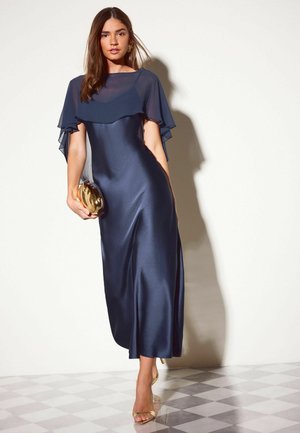 Frau trägt ein marineblaues Satinkleid mit transparenten Umhangärmeln, hält eine goldene Clutch und steht auf einem schachbrettartigen Boden vor einer hellen Wand.