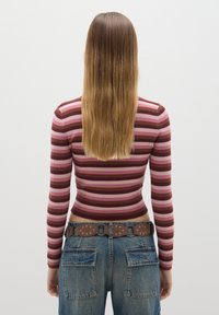 Gestreiftes Langarmtop in Pink, Burgunder und Braun, kombiniert mit blauen Jeans aus Denim, die einen braunen gemusterten Gürtel und abgenutzte Taschen aufweisen.