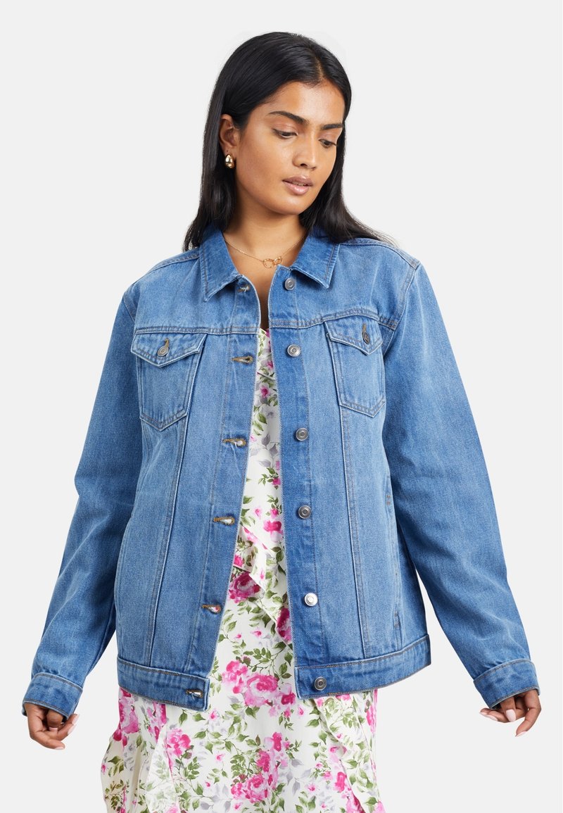 Veste en denim bleu clair avec des fermetures à boutons, deux poches poitrine et une coupe décontractée. Portée par-dessus une robe florale avec des accents roses et verts.