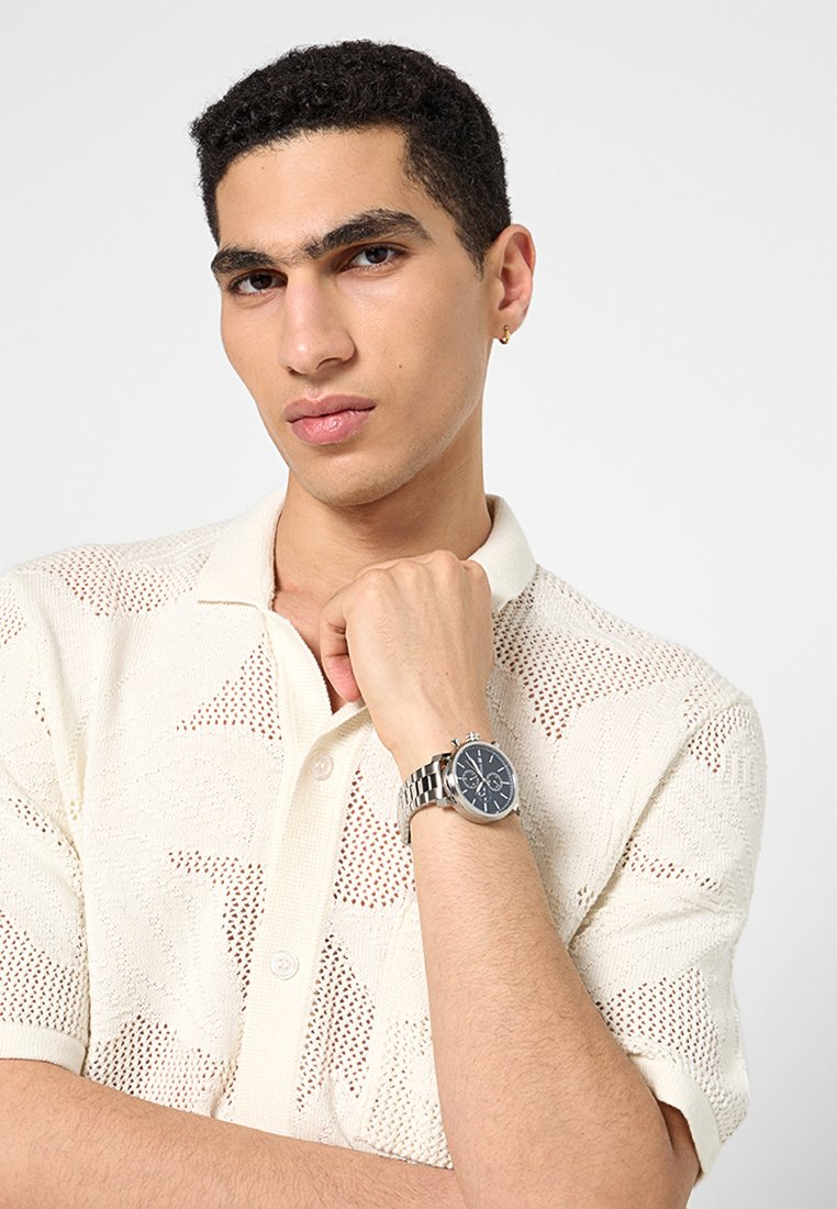 Polo en tricot crème clair à manches courtes, présentant un motif texturé. Le modèle porte une montre argentée avec un cadran sombre.