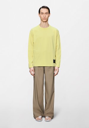 Άτομο που στέκεται, φοράει ανοιχτό κίτρινο μακρυμάνικο πουκάμισο, φαρδιά μπεζ παντελόνια και λευκά σανδάλια τύπου φλιπ φλοπ σε απλό λευκό φόντο.