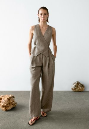 Une personne se tient avec confiance dans une tenue en lin gris, avec un pantalon ample, des sandales rouges et des cheveux lisses, sur un fond minimaliste.