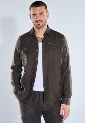 Mann mit kurzen Haaren und Bart, trägt ein aufgeknöpftes dunkelbraunes Hemd über einem weißen Tanktop, steht vor hellem einfarbigem Hintergrund.