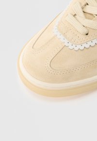 Beżowy zamszowy nosek sneakersa z fakturowanymi kremowymi sznurowadłami, falistą białą lamówką i jasnobrązową gumową podeszwą na białym tle.