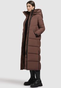 Ilgas, rudas pūkų paltas su gobtuvu, turintis užtrauktuką, šonines kišenes ir siuvinėtą raštą. Derinamas su juodomis nėrimo botais.