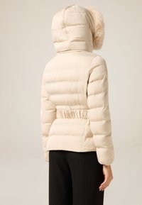 Beżowa pikowana kurtka puchowa z kapturem i sztucznym futerkiem, z detalem marszczonej talii i pikowaną fakturą. Noszona na czarnych spodniach w prążki.