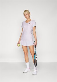 Lys lilla treningsgenser med korte ermer og rund hals, kombinert med matchende shorts. Hvite joggesko og en tenniskølle er avbildet.