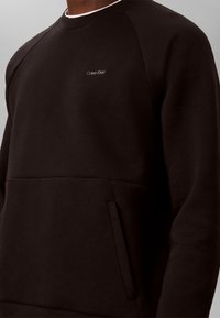 Άτομο φοράει μαύρο φούτερ Calvin Klein με μικρό λογότυπο στο στήθος και μια μπροστινή τσέπη ορατή από το πλάι.
