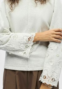 Gros plan d'une personne portant un haut gris clair avec des poignets en dentelle florale brodée et un pantalon marron, les bras croisés doucement.