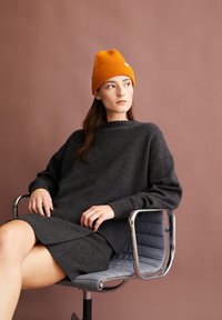 Frau, die eine orange gestrickte Mütze und einen dunkelgrauen Pullover sowie einen Rock trägt, sitzt auf einem grauen Bürostuhl vor einem gedämpften braunen Hintergrund.