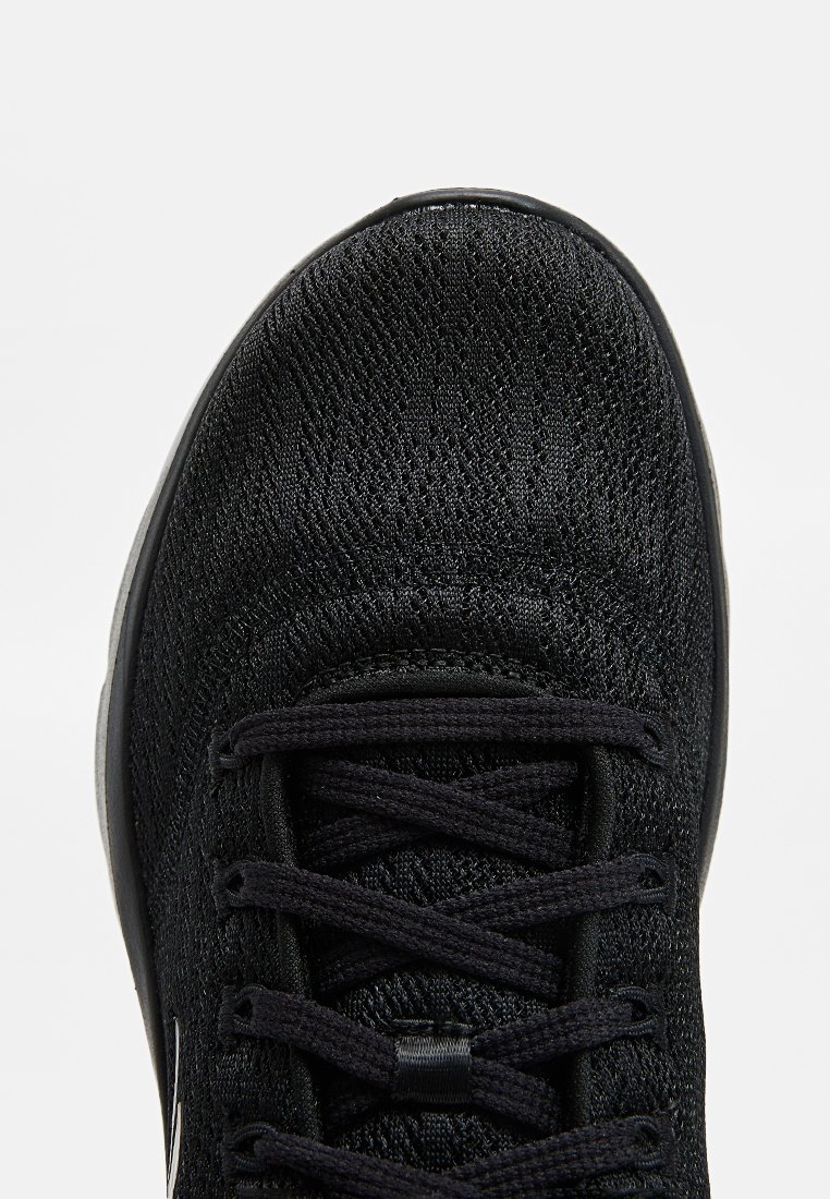 Zapatilla deportiva negra con parte superior de tela texturizada, punta redonda, cordones planos y un logotipo sutil en el lateral. Diseño ligero adecuado para deportes.