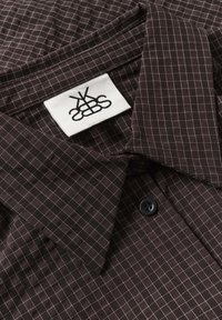 Chemise marron foncé à boutons avec motif quadrillé blanc et bouton noir, avec une étiquette blanche comportant un logo abstrait noir sur le col.