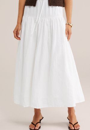 Laisvai krentanti balta midi sijonas su susiaučiuota juosmens linija, derinama su rudos spalvos aukštakulniais sandalais ir auksine apyranke kairėje riešo pusėje.